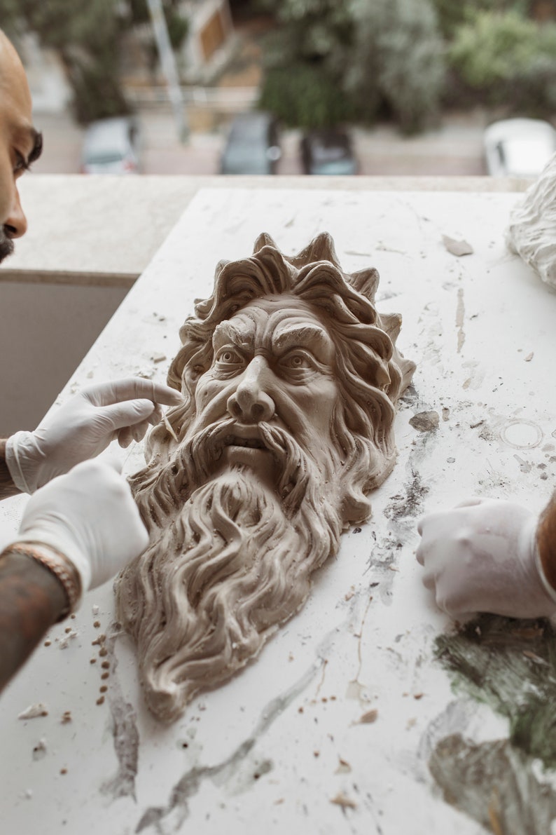 May include: A person is working on a clay sculpture of a man's face with a long beard. The sculpture is on a white surface with some clay debris. The person is wearing white gloves.