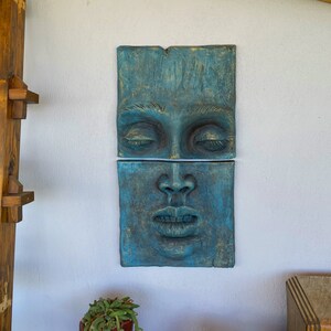 May include: Two-piece wall art featuring a sculpted face with closed eyes and parted lips. The artwork is a teal color with gold accents, mounted on a white wall. The piece is rectangular and has a textured surface.