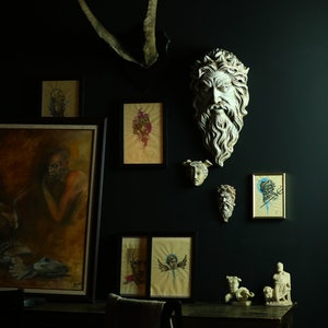 May include: A dark room with a black wall decorated with framed artwork and plaster sculptures. The artwork includes a painting of a man with a beard, a painting of a woman with a crown, and a painting of a woman with wings. The sculptures include a bust of a man with a beard and a bust of a woman with a crown. There is a wooden table with a chair in the foreground.