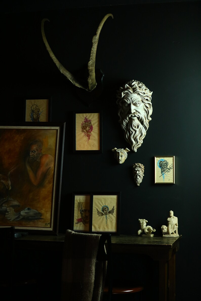 May include: A dark room with a black wall decorated with framed artwork and plaster sculptures. The artwork includes a painting of a man with a beard, a painting of a woman with a crown, and a painting of a woman with wings. The sculptures include a bust of a man with a beard and a bust of a woman with a crown. There is a wooden table with a chair in the foreground.