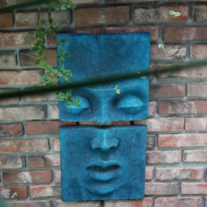 May include: Two teal-colored, square wall sculptures depicting a human face. The top sculpture shows closed eyes, and the bottom one shows a nose and lips. The sculptures are mounted on a brick wall, with greenery in the foreground.