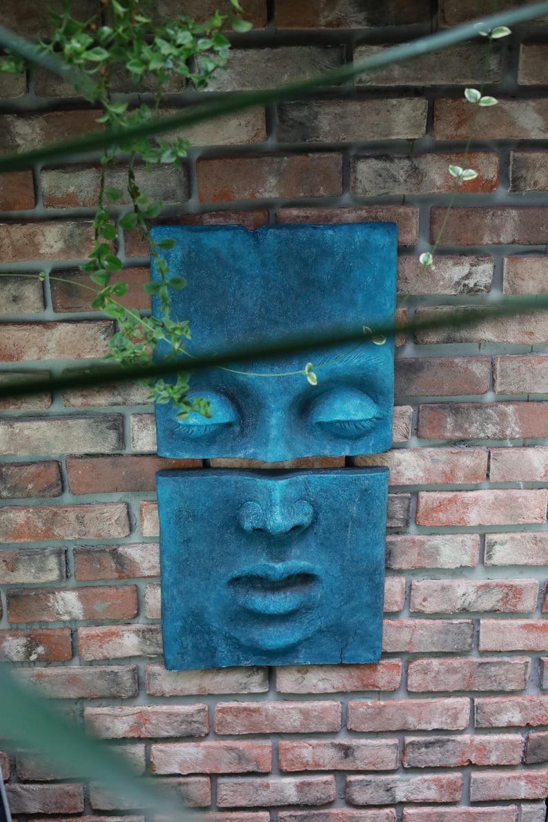 May include: Two teal-colored, square wall sculptures depicting a human face. The top sculpture shows closed eyes, and the bottom one shows a nose and lips. The sculptures are mounted on a brick wall, with greenery in the foreground.