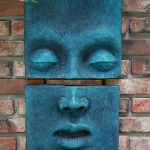 May include: Two teal-colored, rectangular wall sculptures depicting a human face. The top sculpture shows closed eyes, and the bottom shows the nose and lips. The sculptures are mounted on a brick wall.