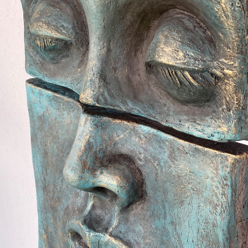 May include: A close-up of a weathered sculpture of a face, split horizontally. The sculpture has a teal and bronze color scheme, with closed eyes and a serene expression. The texture appears rough and aged.