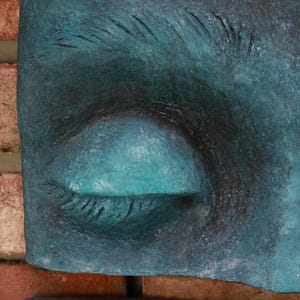 May include: Close-up of a teal-colored sculpture of a closed eye. The eye is set against a backdrop of a brick wall. The sculpture has a textured surface and is painted in shades of teal and black.