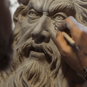 May include: A close-up of a clay sculpture of a man's face with a long beard. The sculpture is being worked on by a hand holding a tool.