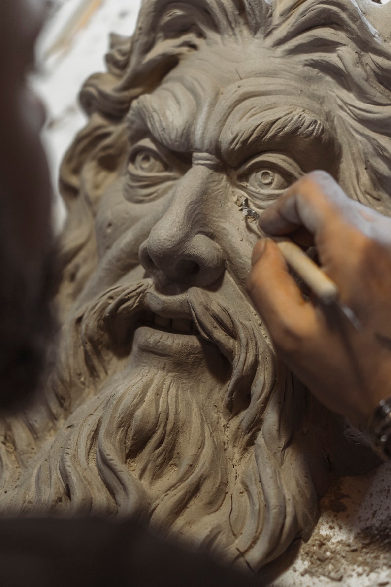 May include: A close-up of a clay sculpture of a man's face with a long beard. The sculpture is being worked on by a hand holding a tool.