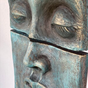 May include: A close-up of a teal and gray sculpture of a face with closed eyes. The sculpture is divided horizontally, with a visible crack. The face has a serene expression, with gold accents highlighting the details.