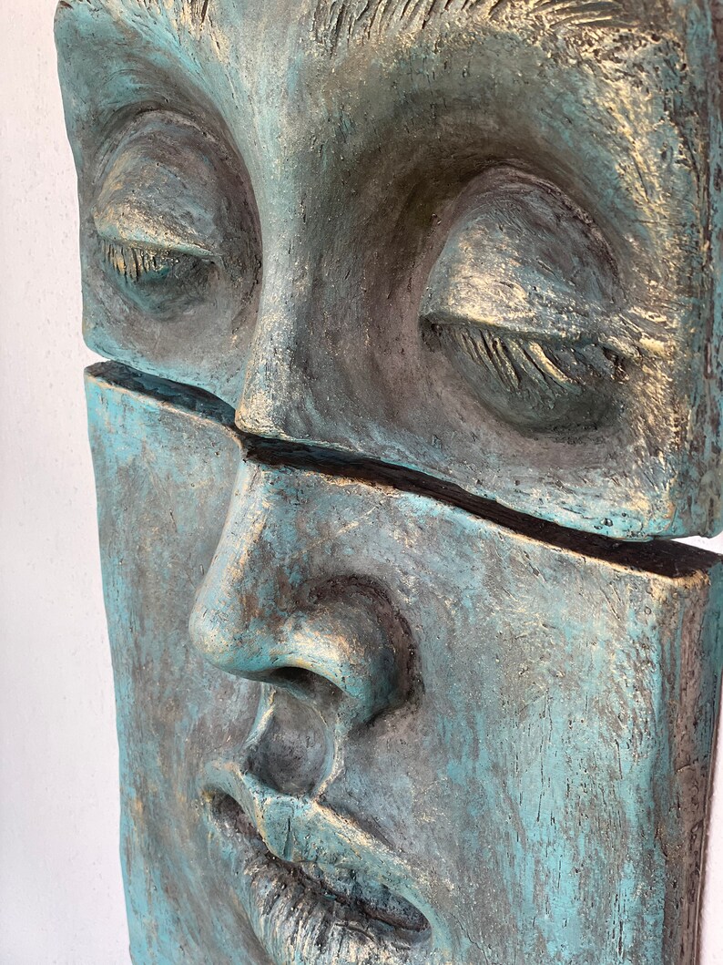 May include: A close-up of a teal and gray sculpture of a face with closed eyes. The sculpture is divided horizontally, with a visible crack. The face has a serene expression, with gold accents highlighting the details.