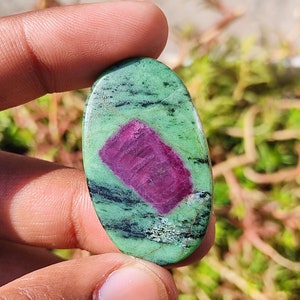 Könnte beinhalten: Ovaler grüner und rosafarbener Edelstein mit schwarzen Adern. Der Edelstein ist im Cabochon-Schliff geschliffen und hat eine glatte, polierte Oberfläche.