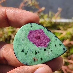 May include: A green and pink gemstone cabochon with a starburst pattern. The gemstone is set in a silver bezel and is ready to be used in jewelry making.
