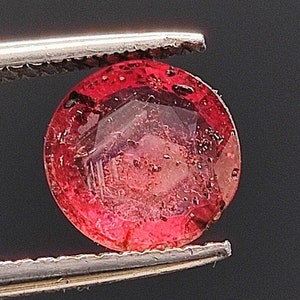 Op de afbeelding: Een ronde, gefacetteerde, rode edelsteen met een licht paarse tint. De edelsteen wordt vastgehouden in een pincet.