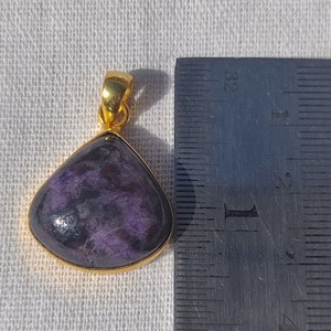Pode incluir: Um pingente de ouro com uma gema roxa e preta. O pingente mede aproximadamente 2,5 cm de comprimento.