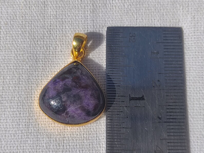 Pode incluir: Um pingente de ouro com uma gema roxa e preta. O pingente mede aproximadamente 2,5 cm de comprimento.