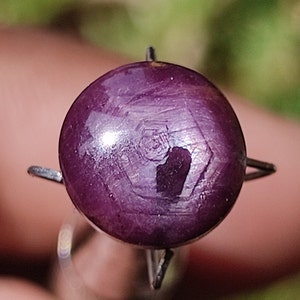 May include: A round, purple gemstone with a star-like pattern in the center. The gemstone is set in a silver prong setting.