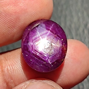 May include: A deep purple star ruby gemstone with a star-shaped reflection in the center. The ruby is cut in a round shape and has a smooth, polished surface.
