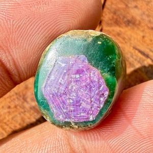 以下が含まれることがあります： 鮮やかな紫色の六角形の模様が中央にあり、濃い緑色のマトリックスに囲まれた、楕円形の磨かれた宝石。 石は指の間に挟まれ、色と質感の自然なバリエーションを示しています。 ユニークなジュエリーです。