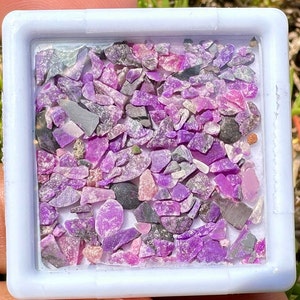 May include: A small white square container filled with a variety of purple and pink gemstone chips. The chips are irregularly shaped and have a sparkly, iridescent finish.