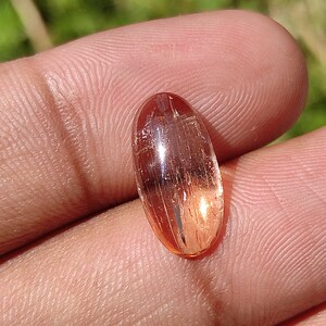 May include: A translucent, oval-shaped gemstone with a reddish-orange hue and visible internal striations. The gemstone is set in a cabochon cut.