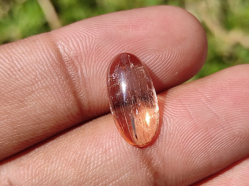 May include: A translucent, oval-shaped gemstone with a reddish-orange hue and visible internal striations. The gemstone is set in a cabochon cut.