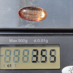 May include: A close-up of a digital scale displaying "0.000 g" with a light pink oval-shaped gemstone on top of the scale.