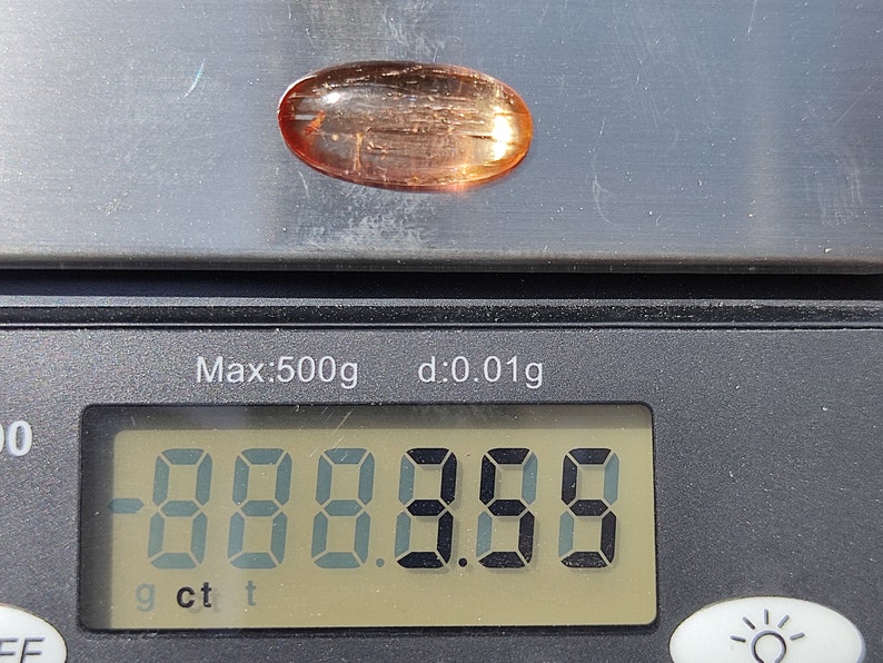 May include: A close-up of a digital scale displaying "0.000 g" with a light pink oval-shaped gemstone on top of the scale.