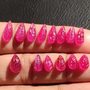 Op de afbeelding: Een verzameling van 14 roze robijn edelsteen kralen, gesneden in een blaadjesvorm. De kralen zijn in twee rijen op een hand gerangschikt.