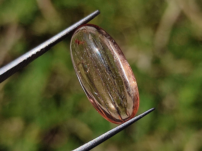 May include: A translucent, oval-shaped gemstone with a pink hue and visible internal striations. The gemstone is held by tweezers against a green background.