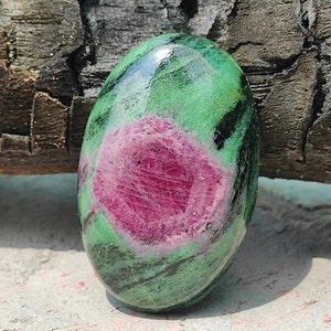 Könnte beinhalten: Ein ovaler Edelstein in Grün- und Rosatönen mit schwarzen Streifen. Der Edelstein ist glatt und poliert.