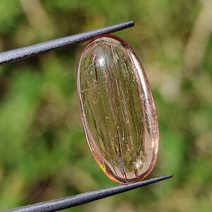May include: A translucent, oval-shaped, light pink gemstone with visible striations. The gemstone is held by a pair of tweezers.