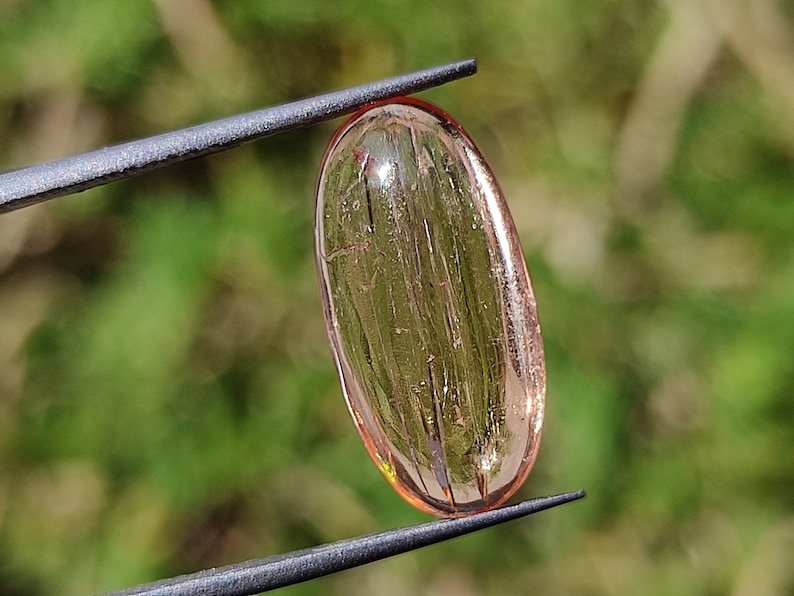 May include: A translucent, oval-shaped, light pink gemstone with visible striations. The gemstone is held by a pair of tweezers.