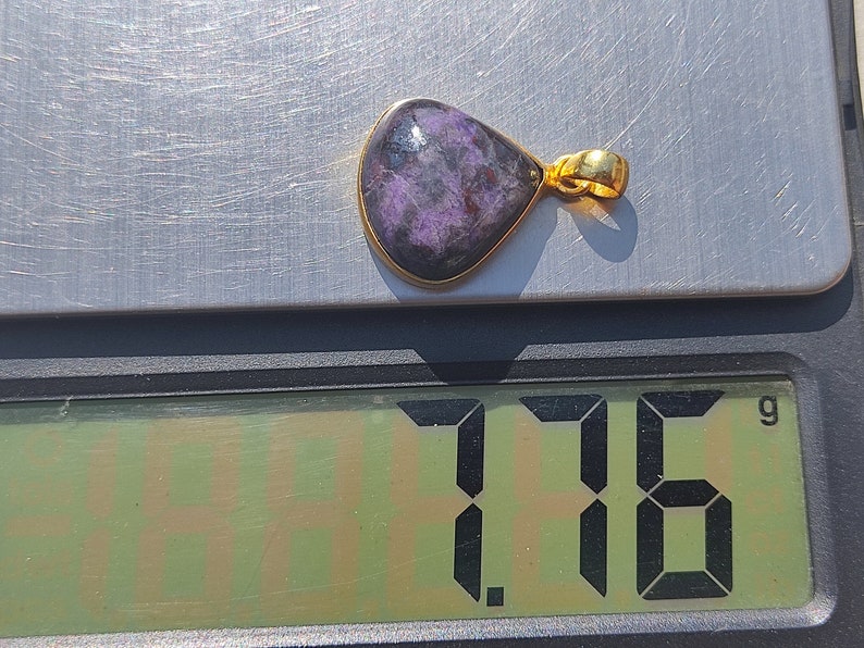 Pode incluir: Um pingente roxo e dourado com uma pedra em forma de l&aacute;grima. O pingente pesa 1,76 gramas.