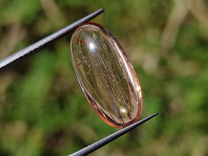 May include: A translucent, oval-shaped, light pink gemstone with visible internal striations. The gemstone is held by tweezers against a blurred green background.