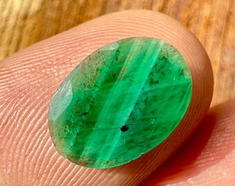 May include: Oval-shaped, faceted emerald gemstone. The gem displays a vibrant green color with internal veining and a small black inclusion. The stone is held between fingers, showcasing its polished surface and clarity.