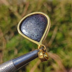 Pode incluir: Um pingente de ouro com uma gema cinza escura engastada em forma de l&aacute;grima. O pingente tem um aro para prender a uma corrente.