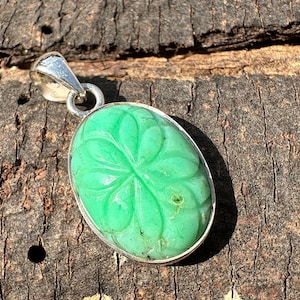 Pode incluir: Um pingente de pedra preciosa verde talhada com uma argola de prata. A pedra preciosa é de forma oval e tem um desenho floral talhado.