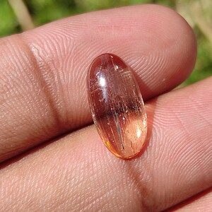 May include: A translucent, oval-shaped gemstone with a reddish-brown hue and visible internal striations. The gemstone is set in a cabochon cut.