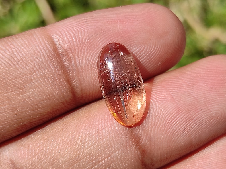 May include: A translucent, oval-shaped gemstone with a reddish-brown hue and visible internal striations. The gemstone is set in a cabochon cut.