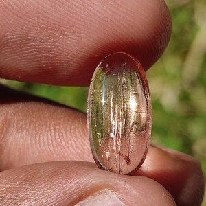 May include: A pink and gold oval-shaped gemstone with a striated pattern. The gemstone is held in a hand.