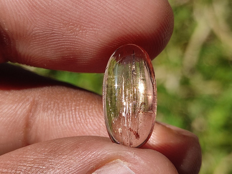 May include: A pink and gold oval-shaped gemstone with a striated pattern. The gemstone is held in a hand.
