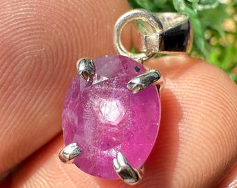 Puede incluir: Un colgante de rubí ovalado y facetado con una fianza de plata. El rubí es de color rosa intenso y está sujeto por cuatro garras de plata. El colgante está en la mano de una persona.