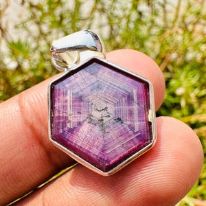 May include: A hexagonal pendant featuring a deep red gemstone. The gemstone displays a distinctive star-shaped pattern within its polished surface. The pendant is set in a silver metal frame and has a silver bail for a chain.