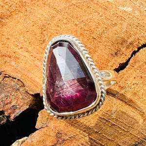 Op de afbeelding: Een zilveren ring met een grote, gefacetteerde, paarse edelsteen gezet in een band met een gedraaid touwontwerp.