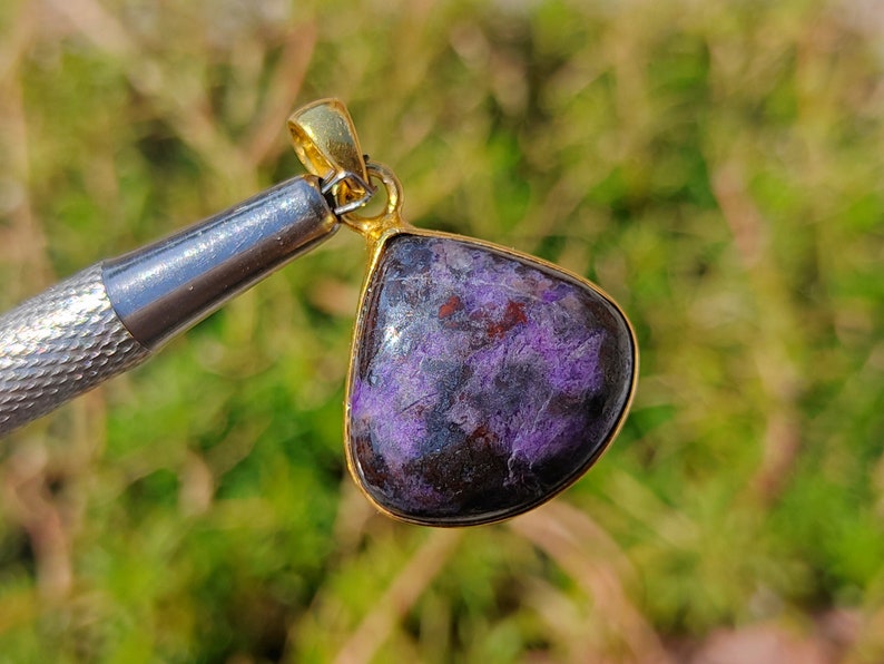 Pode incluir: Um pingente em forma de l&aacute;grima com uma gema roxa e marrom engastada em uma moldura de metal dourado.