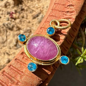 May include: A gold pendant with a pink oval gemstone surrounded by four smaller blue gemstones. The pendant has a gold loop at the top for attaching to a chain.