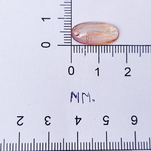 May include: A light brown oval-shaped gemstone measuring approximately 1.5 cm in length. The gemstone is sitting on a white surface with a ruler for scale.