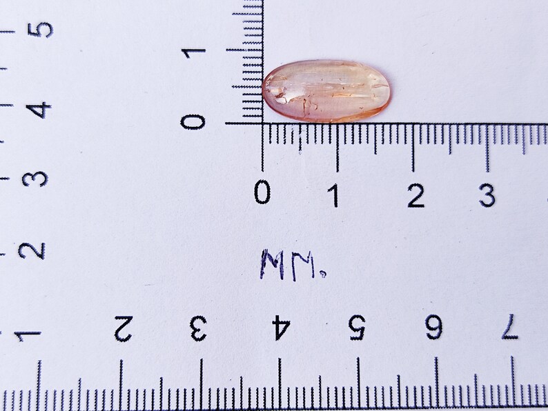 May include: A light brown oval-shaped gemstone measuring approximately 1.5 cm in length. The gemstone is sitting on a white surface with a ruler for scale.