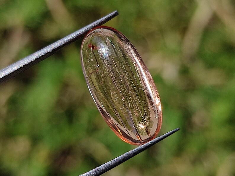 May include: A translucent, oval-shaped, light pink gemstone with visible internal lines. The gemstone is held by tweezers against a blurred green background.