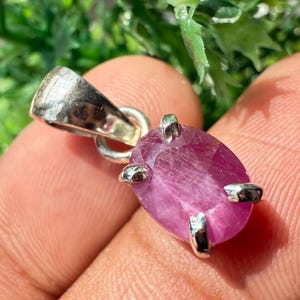 Puede incluir: Primer plano de un colgante de rubí. El colgante presenta una gran piedra preciosa de color rosa oscuro, de forma ovalada, engastada en un marco de metal plateado. La argolla plateada está unida a la parte superior del colgante.
