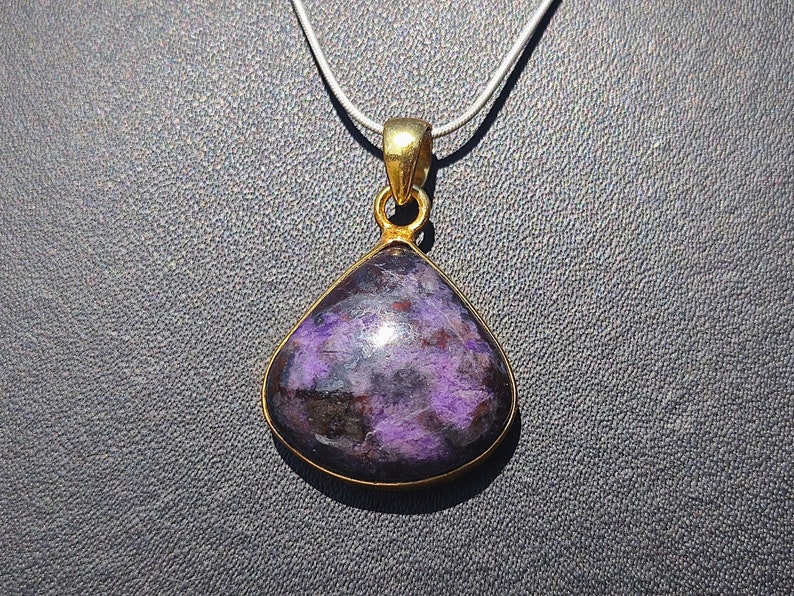 Pode incluir: Um pingente de ouro com uma gema roxa e preta. O pingente tem a forma de uma l&aacute;grima e tem uma fivela de ouro.